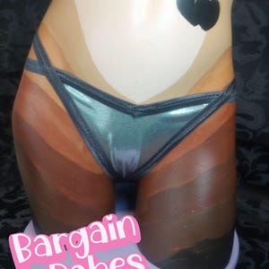 May include: A metallic teal bikini bottom with thin, crisscrossing side straps displayed on a mannequin. The garment features a smooth, shiny finish and a V-shaped front. A black heart-shaped graphic is visible on the upper hip area, and the text Bargain Babes is printed in white and pink at the bottom.