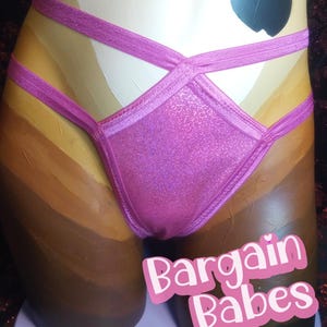 Puede incluir: Una braguita de bikini rosa brillante con tirantes entrecruzados. El bikini se muestra en un maniquí con un degradado de tonos de piel. El texto "Bargain Babes" se muestra en rosa y blanco.