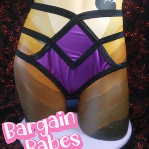 May include: A purple and black lingerie set with a geometric design. The set features a diamond-shaped front panel and black straps. The words "Bargain Babes" are in pink text at the bottom.