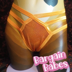 Puede incluir: Lencería naranja con un panel frontal brillante y correas elásticas. La prenda se exhibe en un maniquí con un degradado de color marrón y beige. El texto "Bargain Babes" se muestra en la parte inferior.