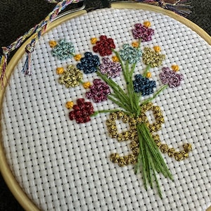 May include: A cross-stitch embroidery of a bouquet of flowers in a wooden hoop. The flowers are in shades of red, blue, green, yellow, and purple. The stems are green, and the bouquet is tied with a gold ribbon. The hoop is 6 inches in diameter.