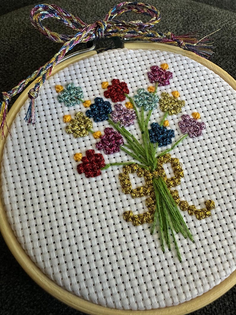 May include: A cross-stitch embroidery of a bouquet of flowers in a wooden hoop. The flowers are in shades of red, blue, green, yellow, and purple. The stems are green, and the bouquet is tied with a gold ribbon. The hoop is 6 inches in diameter.