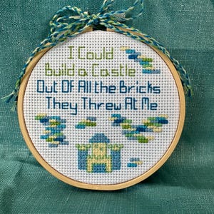 May include: A cross-stitch embroidery hoop with the text "I Could Build a Castle Out Of All the Bricks They Threw At Me." The design features a castle and brick patterns in blue, green, and yellow. The hoop is trimmed with a blue and green string.
