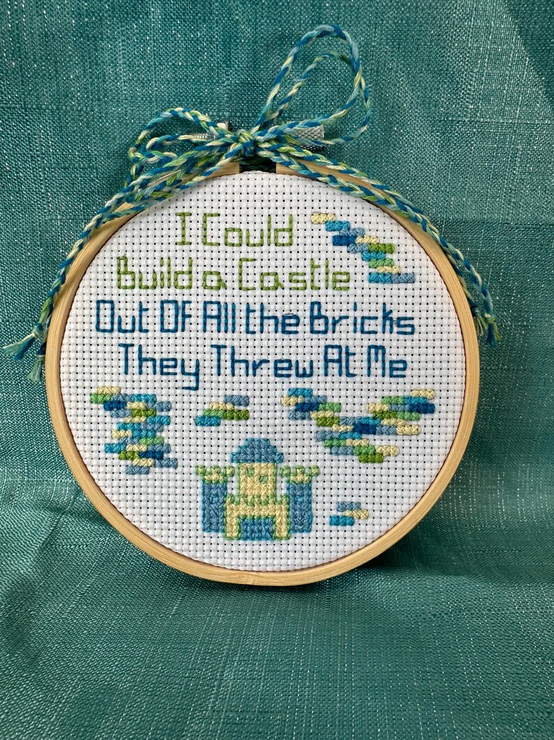 May include: A cross-stitch embroidery hoop with the text "I Could Build a Castle Out Of All the Bricks They Threw At Me." The design features a castle and brick patterns in blue, green, and yellow. The hoop is trimmed with a blue and green string.