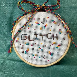 May include: A round cross-stitch embroidery hoop with the word "GLITCH" stitched in dark brown. The design includes colorful cross-stitch accents in teal, yellow, and maroon. A multi-colored braided cord forms a bow at the top.