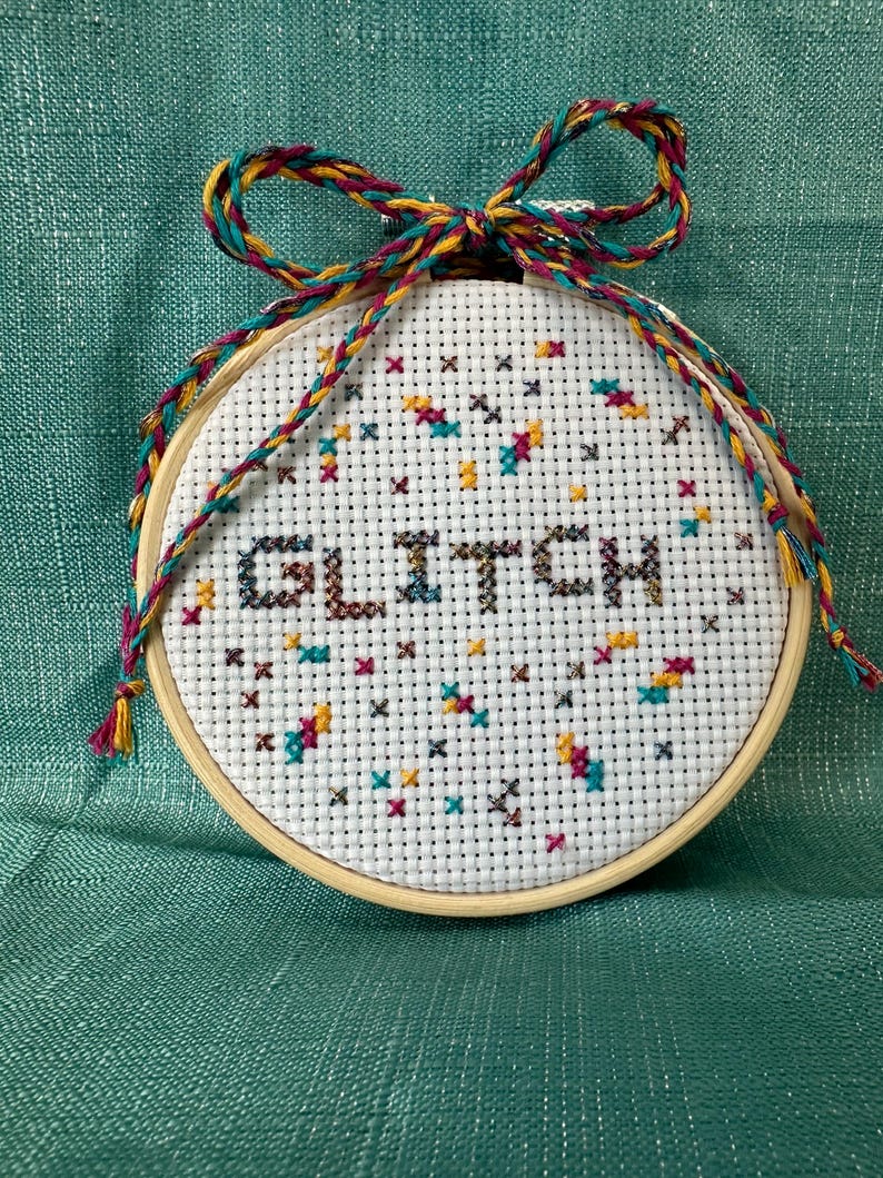 May include: A round cross-stitch embroidery hoop with the word "GLITCH" stitched in dark brown. The design includes colorful cross-stitch accents in teal, yellow, and maroon. A multi-colored braided cord forms a bow at the top.