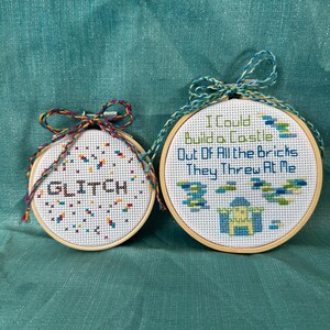 May include: Two cross-stitch hoops with wooden frames. One hoop says "GLITCH" in black and white, with colorful accents. The other says "I Could Build a Castle Out Of All the Bricks They Threw At Me" with a castle design.