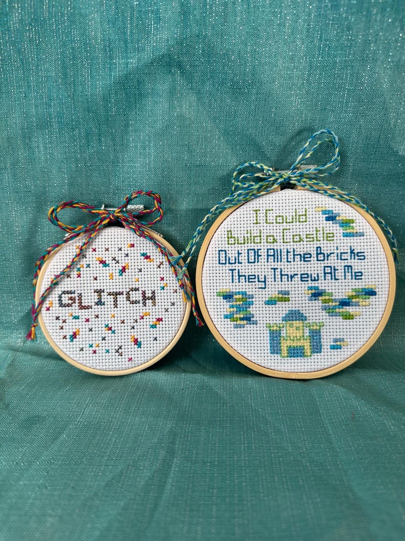 May include: Two cross-stitch hoops with wooden frames. One hoop says "GLITCH" in black and white, with colorful accents. The other says "I Could Build a Castle Out Of All the Bricks They Threw At Me" with a castle design.