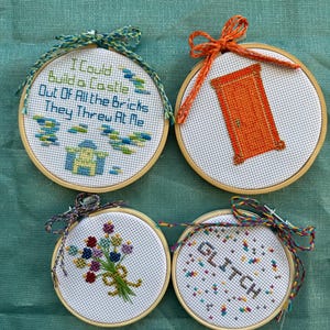 May include: Four round cross-stitch hoops with various designs. One hoop features the text "I Could Build a Castle Out Of All the Bricks They Threw At Me." Other hoops display a door, a bouquet of flowers, and the word "GLITCH."