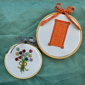 May include: Two small, round cross-stitch hoops. One features an orange door design, the other a bouquet of flowers. Both are set against a teal background. The hoops are framed in wood and have decorative bows.