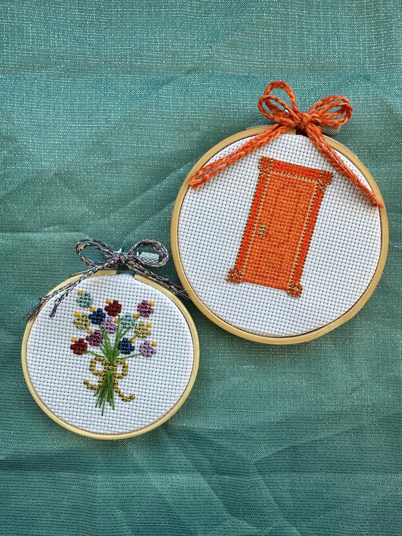 May include: Two small, round cross-stitch hoops. One features an orange door design, the other a bouquet of flowers. Both are set against a teal background. The hoops are framed in wood and have decorative bows.