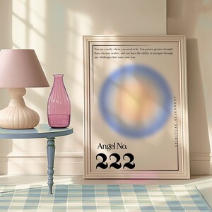 May include: A framed poster with a blurred image of a circle in the center. The poster has the text "Angel No. 222" and "Spiritual Alignment" on it. The poster is sitting on a table with a pink lamp and a pink glass vase.
