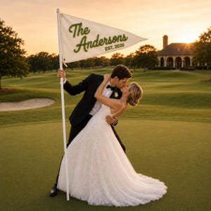 May include: A triangular white pennant flag attached to a white pole, featuring the green embroidered text The Andersons and EST. 2026. The flag is made of fabric with metal grommets and is held upright by a person in a black suit and a white dress.