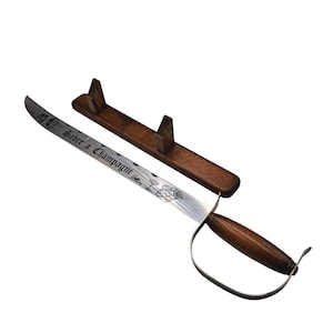 May include: A champagne saber with a wooden handle and a silver blade, resting on a wooden stand. The blade is engraved with the words "Sabre a Champagne" and floral designs. The handle is dark brown.