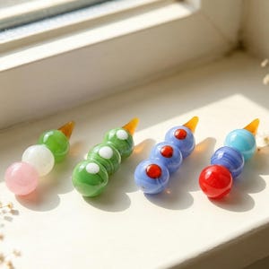 Pode incluir: Quatro ornamentos de vidro em estilo dango, cada um consistindo em três contas esféricas empilhadas verticalmente e cobertas com uma pequena ponta de cor âmbar. As contas apresentam várias cores, incluindo rosa, branco, verde, azul e vermelho com alguns padrões marmorizados.