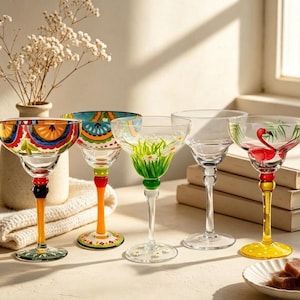 May include: Five glass margarita glasses with distinct designs. One features orange and blue patterns, another has green grass and white daisies, and one displays a pink flamingo with green palm leaves. The stems are colored in yellow, orange, and green.