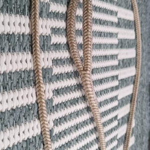 Puede incluir: Cuerda trenzada beige con un lazo, posiblemente un asa o elemento decorativo. La cuerda está sobre una superficie texturizada a rayas grises y blancas. El extremo de la cuerda tiene una borla y un acento verde.