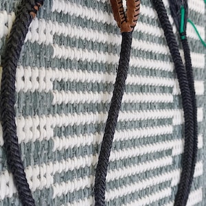 Puede incluir: Un látigo trenzado gris oscuro con un mango de cuero marrón y un lazo en el extremo. El látigo está sobre una superficie a rayas blancas y grises.