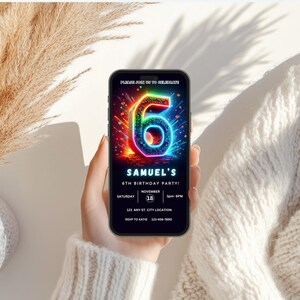 May include: A mobile phone screen shows a colourful birthday invitation. The design includes a neon number 6, the text "SAMUEL'S 6TH BIRTHDAY PARTY!" and event details. The phone is held against a soft, textured background.