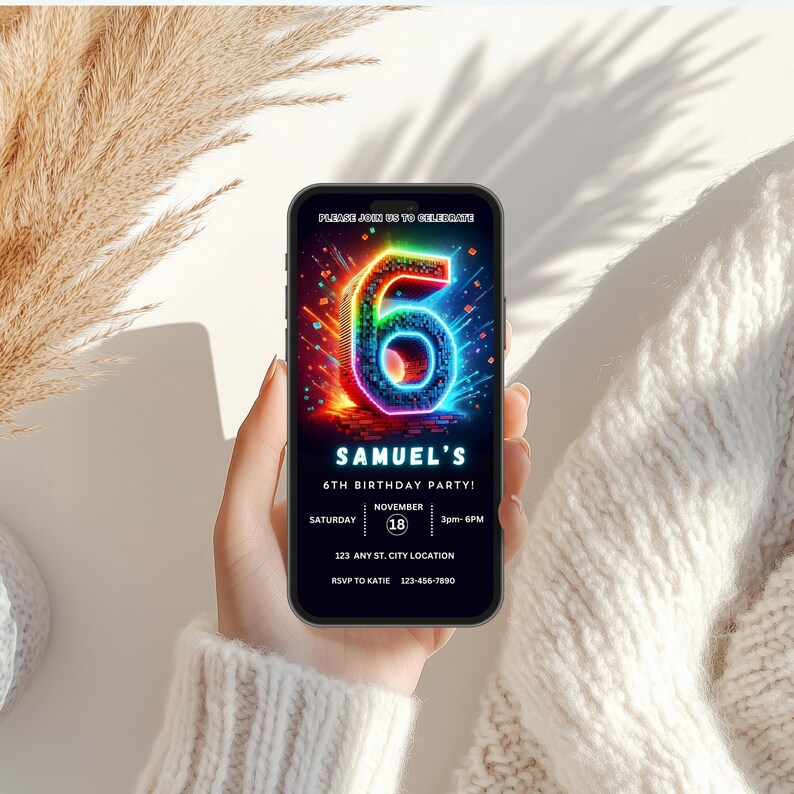 May include: A mobile phone screen shows a colourful birthday invitation. The design includes a neon number 6, the text "SAMUEL'S 6TH BIRTHDAY PARTY!" and event details. The phone is held against a soft, textured background.