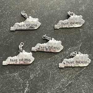 May include: Five silver-toned metal charms shaped like the state of Kentucky, each featuring a textured surface and the stamped text "BLUE GRASS" in capital letters. Each charm includes a small metal jump ring attached to the top for hanging.
