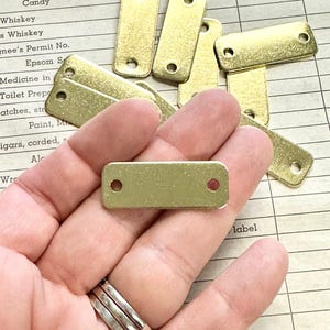 May include: A hand holding a group of small, rectangular, gold-coloured metal tags with two holes in each. The tags are on a white surface with black lines.