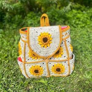 May include: A handmade crochet backpack featuring a sunflower design. The bag is primarily off-white with yellow and brown sunflower appliqués. It has a top handle and adjustable straps. The backpack is made of yarn and has a button closure.