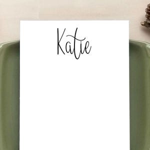 May include: A white notepad with the name "Katie" in elegant black script. The notepad is placed on a green ceramic dish, with pinecones, pencils, and twine adding a natural aesthetic.