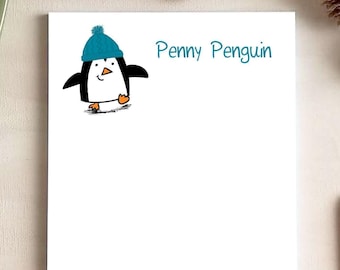 May include: A white rectangular notepad featuring a cartoon penguin wearing a blue knit hat and the text Penny Penguin in blue script. The penguin has a black and white body with orange feet and a small orange beak, positioned at the top left.