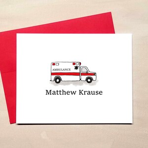 May include: A white card with a red envelope behind it. The card features a hand-drawn ambulance with the word "AMBULANCE" above the vehicle and the name "Matthew Krause" below it. The ambulance is red and white.