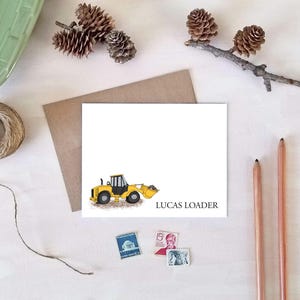 May include: A white card with a yellow and black loader illustration and the text "LUCAS LOADER". The card is surrounded by decorative elements including pinecones, pencils, and stamps, suggesting a stationery or gift theme.