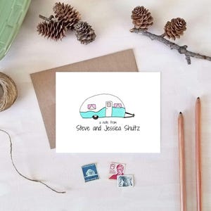 May include: A white card with a teal and white camper illustration and the text "a note from Steve and Jessica Shultz." The card is on a white surface with a brown envelope, pencils, stamps, twine, and pinecones.