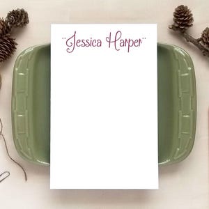 May include: White notepad with maroon cursive text 'Jessica Harper' at the top. The notepad is displayed on a sage green rectangular dish.  Pine cones and pencils are arranged around the dish on a light beige surface. The notepad is blank below the name, ready for writing. This stationery item is perfect for personal use or as a gift.