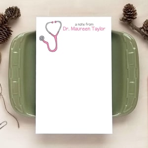 May include: A white rectangular notepad featuring a pink stethoscope illustration and the printed text a note from Dr. Maureen Taylor in pink and black ink. The notepad is positioned vertically on a solid surface for writing notes.