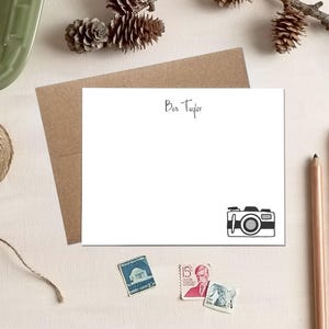 May include: White stationery with a black camera illustration and the name "Ben Taylor" in script. Includes a brown envelope, two pencils, and two postage stamps. The stationery is on a light wood surface.