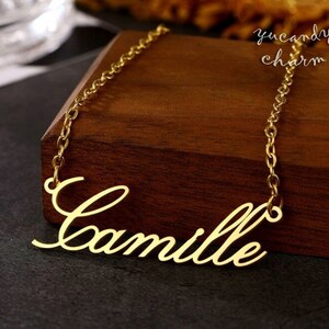 May include: A gold necklace with a cursive nameplate pendant that reads "Camille". The necklace is on a wooden surface.