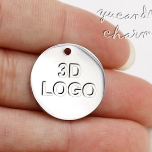 Puede incluir: Un charm redondo plateado con un agujero en la parte superior. El charm tiene las palabras "3D LOGO" grabadas.