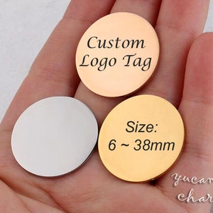 以下が含まれることがあります： 異なる色の3つの丸い金属タグ。金のタグには「サイズ：6〜38mm」と書いてあります。銀のタグは空白です。銅のタグには「カスタムロゴタグ」と書いてあります。