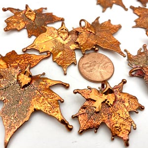May include: Copper-colored maple leaf charms with textured surfaces and small loops for hanging. The leaves are in various stages of detail, with some showing intricate vein patterns. A penny is included for size reference.