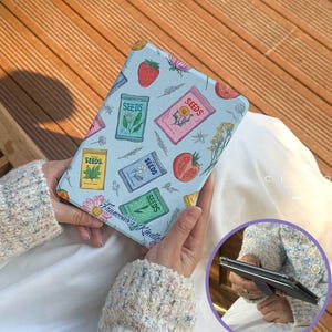 Op de afbeelding: Een lichtblauwe Kindle-hoes met een herhalend patroon van zaadzakjes, aardbeien en tomaten. De hoes heeft de tekst "Francesco's Kindle" en wordt in de handen van een persoon gehouden.