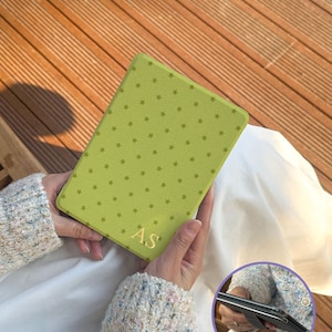 May include: A lime green e-reader cover featuring a repeating pattern of small dark green stars and the gold-colored initials AS embossed at the bottom. The cover is shown in a closed position and also from a side angle in a circular inset.