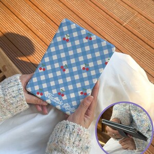 Puede incluir: Una funda Kindle a cuadros azules y blancos con ilustraciones de cerezas rojas. La funda tiene el texto "Francesca's Kindle" en una fuente cursiva. La funda está sujeta en las manos de una persona.