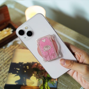 May include: A white smartphone with a pink pop-up grip featuring a floral design. The grip has a pink background with a white rose and a heart. The phone is held by a person.