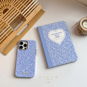 Puede incluir: Una funda de teléfono azul y blanca con la palabra "Cisco" impresa en ella. La funda tiene un diseño que se asemeja a un cuaderno de composición. Un cuaderno de composición azul y blanco con un corazón y las palabras "Composition Book Cisco" impresas en él.