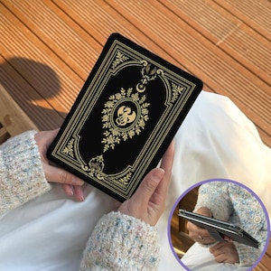 May include: Black tablet case featuring a gold floral and dragon motif. The rectangular case has rounded corners. The design incorporates a dragon, roses, and a crescent moon. The case is held in a person's hands.