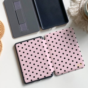 Puede incluir: Una funda para lector electr&oacute;nico de color rosa claro con un patr&oacute;n de puntos negros y las letras doradas AS impresas en la portada. La funda se muestra abierta, revelando un interior gris oscuro con correa, y otra funda cerrada.