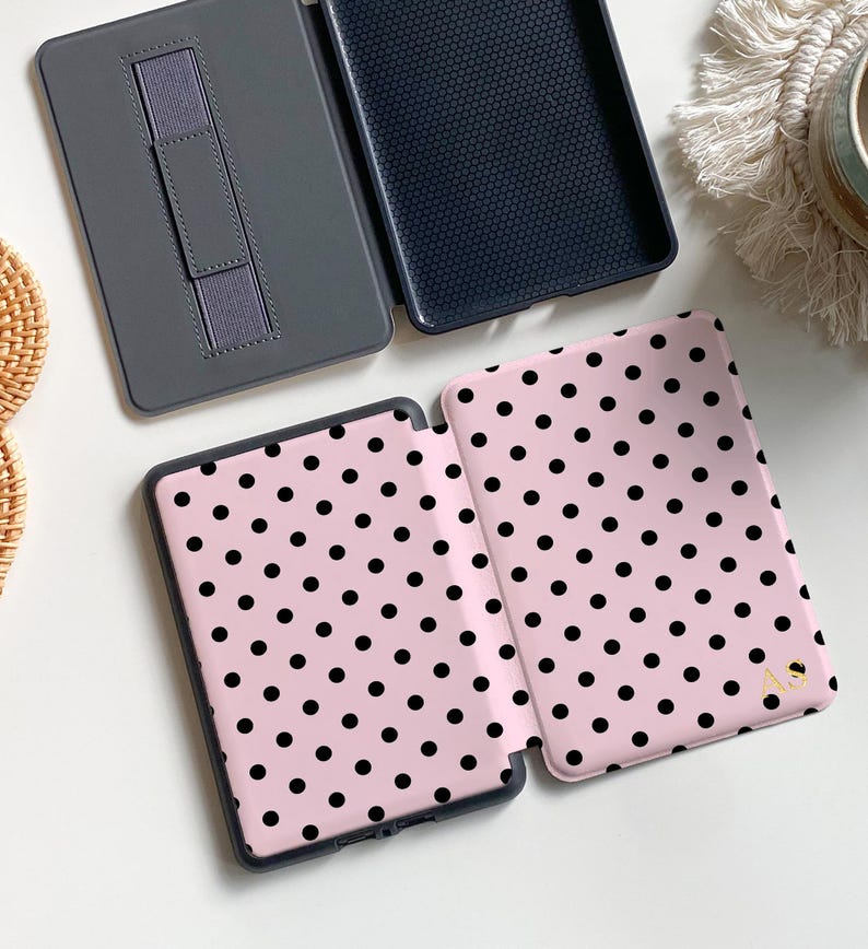 Puede incluir: Una funda para lector electr&oacute;nico de color rosa claro con un patr&oacute;n de puntos negros y las letras doradas AS impresas en la portada. La funda se muestra abierta, revelando un interior gris oscuro con correa, y otra funda cerrada.