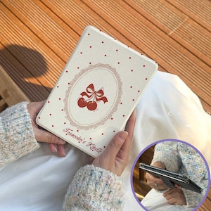 Op de afbeelding: Crèmekleurige Kindle-hoes met een rood kersendesign en de tekst "Francesca's Kindle". De hoes heeft een stippenpatroon en een ovale lijst rond de kersen. De hoes is rechthoekig en heeft een magnetische sluiting.