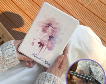 May include: A cream Kindle case with a watercolour design of pink cherry blossoms. The case has the text "Francesco's Kindle" in a script font. The case is held open, revealing the device inside.