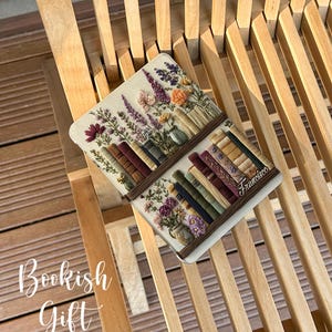 May include: A decorative book-themed item featuring embroidered flowers and books. The item is rectangular with a light-colored background and a brown border. The text "Bookish Gift" is visible in the image.